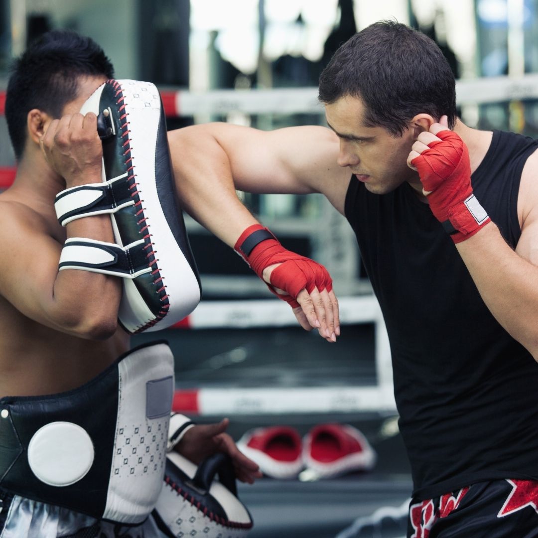 Image of two muay thai guys training with one holding the muay thai pads and the other throwing an elbow at the muay thai pads