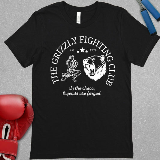 Black shirt with a white design. The design has the words The Grizzly Fighting Club and has a Muay Thai guy doing the Wai Khru next to a Grizzly bear. At the bottom of the design has the words In the Chaos, legends are forged.