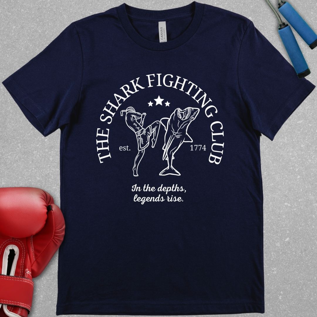 Navy shirt with the slogan The Shark Fighting Club . It has a picture of a Muay Thai fighter throwing a knee at the shark with the phrase In the depths, legends will rise.