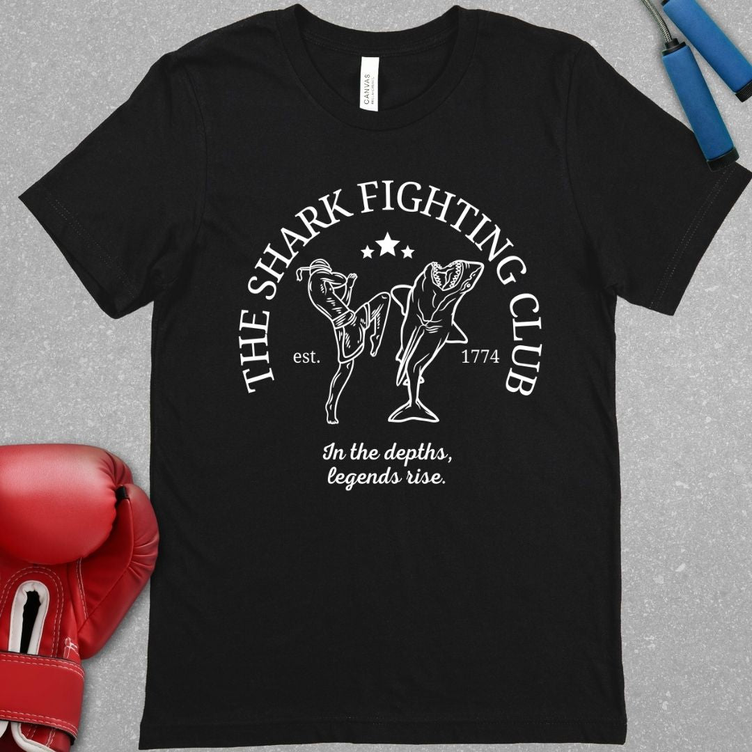 Black shirt with the slogan The Shark Fighting Club. It has a picture of a Muay Thai Fighter throwing a knee at the shark with the phrase In the depths, legends will rise.