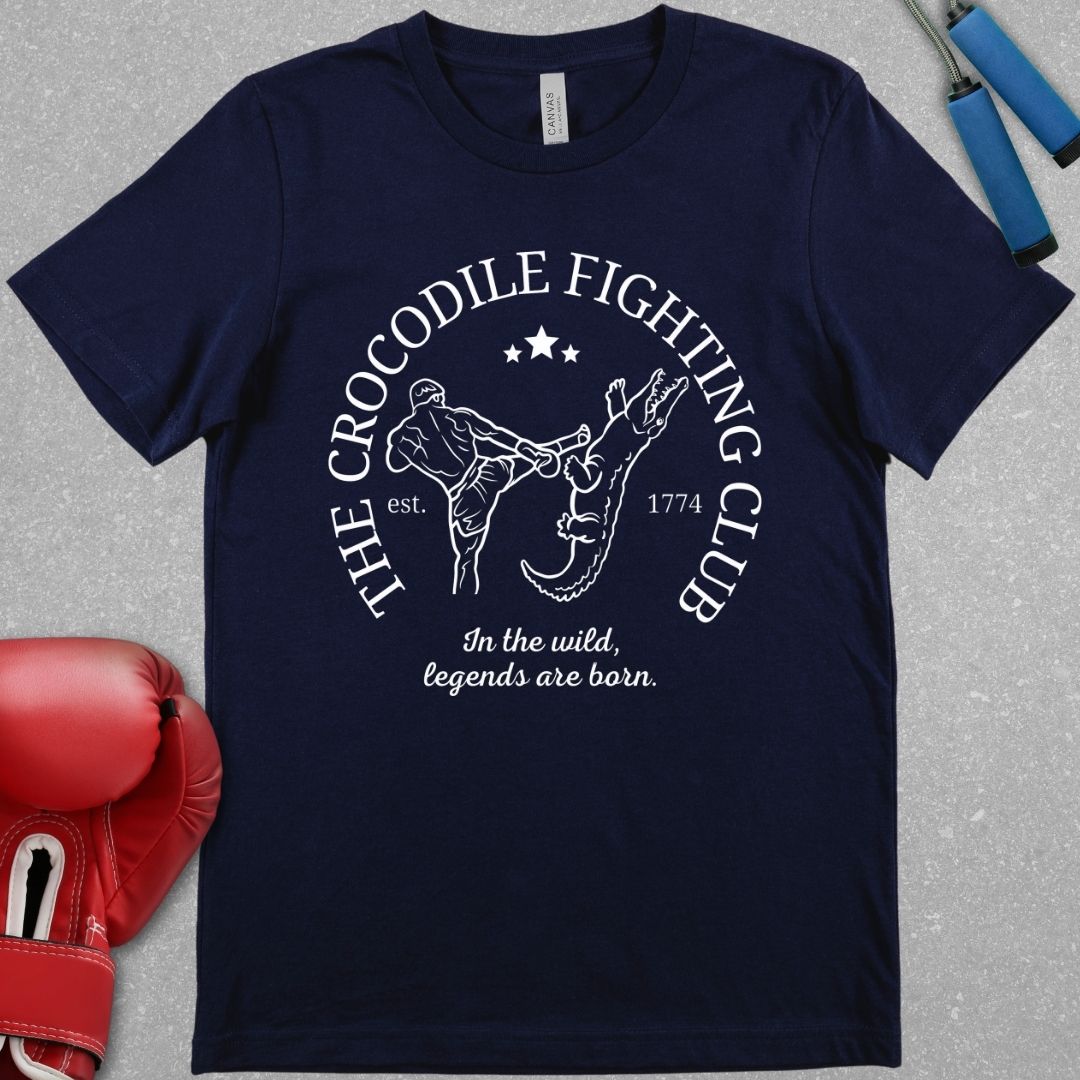 Navy shirt with a white design. The design has the words The Crocodile Fighting Club and has a Muay Thai guy throwing a kick to a crocodile's head. At the bottom of the design has the words In the wild, legends are born.